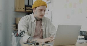 Young employee making online video call with laptop speaking and waving hand in office. Man with tattoo and piercing wearing informal clothing discussing work.