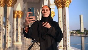 Woman in takes selfie pic in front of mosque in UAE spbas. Young female holding smartphone in hand and taking photo, looking with happy smile and standing on background of white architecture outdoors