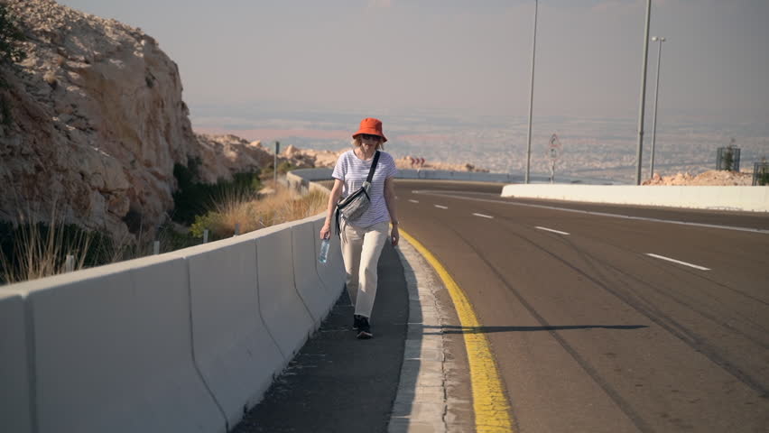 Woman traveler walks on road and drinks water outdoors spbas. Beautiful female walking along highway and looking with smile, drinking aqua and resting after trip on summer day. Pretty lady strolls in