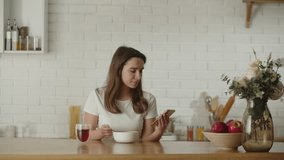 Video of a woman eating oatmeal for breakfast while sitting in the kitchen looking at something on her phone. A brunette is watching funny videos on her phone while having breakfast.