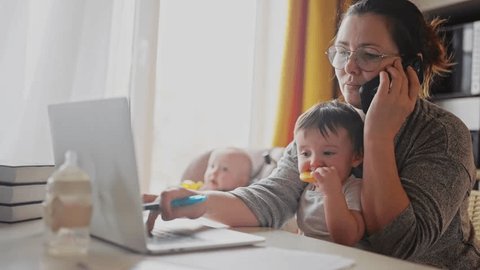 mother working from home remotely with baby daughter in his arms. pandemic remote work business concept. mother tries to work at home in fun kitchen, baby children interfere sitting on their hands – Stockvideo