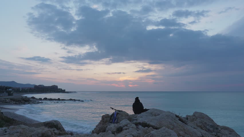 time lapse captures sun rise over lone man meditating on rocks looking into sea and clouds