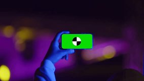 Male hand holds phone with chromakey over his head, shooting video at party, mockup. Concept of concert, party and holiday in neon color