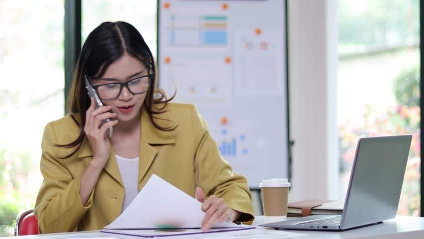 A businesswoman on the phone calling a client is reading some documents on her desk in the office. Woman talking on the phone to someone while analyzing documents and reading monthly work schedule. | Shutterstock HD Video #1101230591