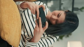 Cheerful Young beautiful Asian woman gamer holding smartphone in hand playing mobile MMORPG MOBA game get victory Express happy Won Joyful Excited Face sit on couch in Living Room. Vertical Video Shot