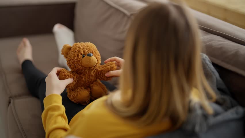 Back view child sitting on couch at home with teddy bear in hands. Girl with broken leg with white plaster cast. Recovery and treatment at home after injury. Support sad children with problem health.