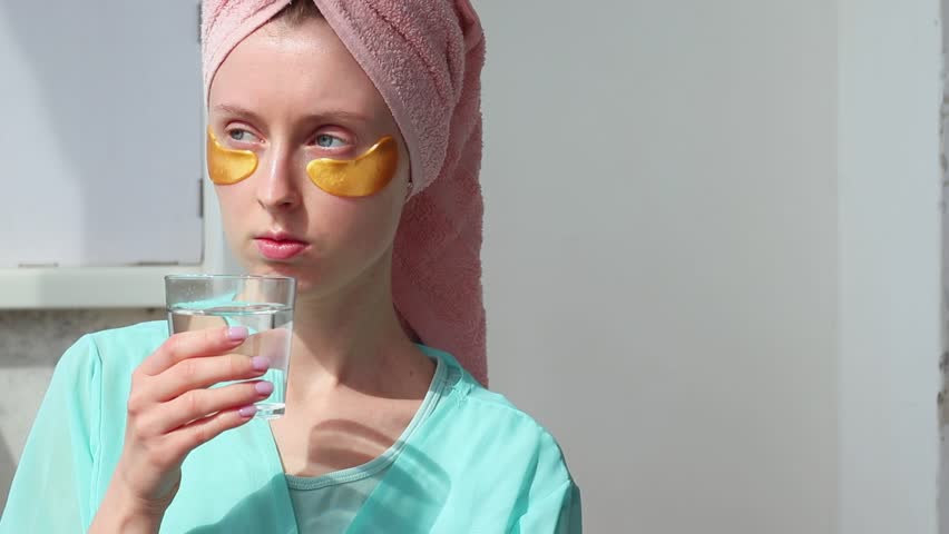 Young girl bathrobe in a spa salon puts gold cosmetic patches on her eyes. A girl with a towel on her head looks in the mirror. Woman combs her wet hair after a shower and drinking a water morning