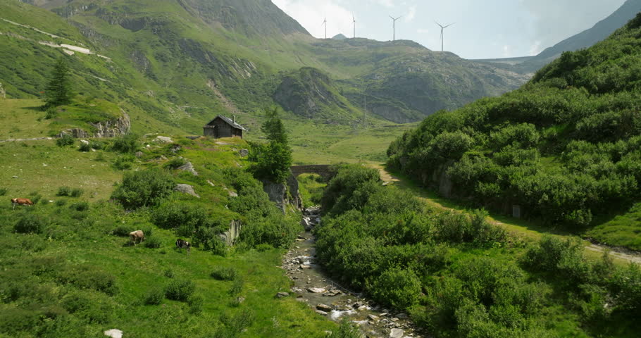 Green meadow with lots of vegetation and shot under an old stone bridge, wind power windmills on the horizon. grimselpass, switzerland. Aerial shot.