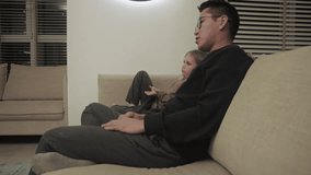 Side view of father and daughter sitting on couch and watching television while staying at home, in a modern apartment at the evening. Father explains something to his daughter during to watching TV