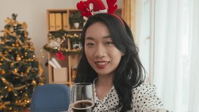 Asian young woman in xmas headband sitting in front of camera in home office, pronouncing Christmas toast and having some wine while communicating in video chat