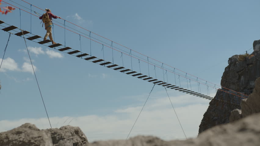 Woman tourist walks along suspension bridge in mountains against background of blue sky.