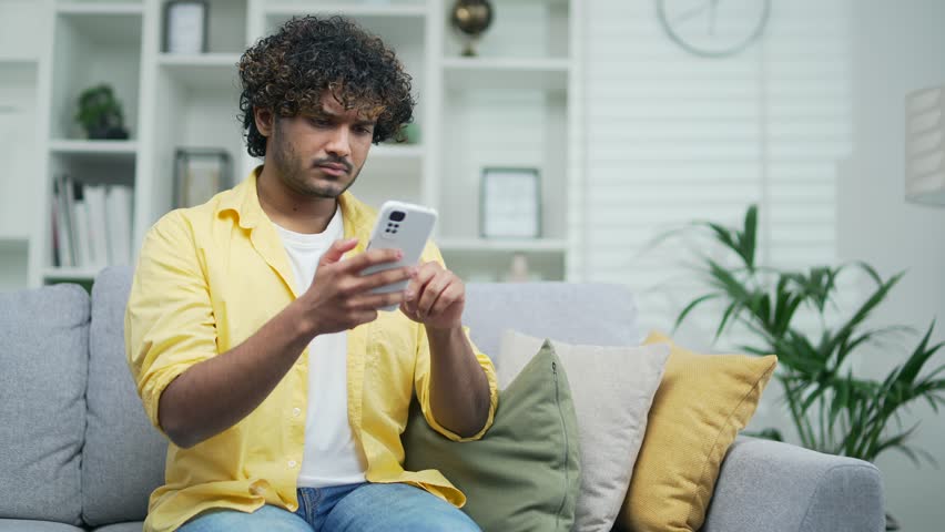 Young man receives a notification with a bad news while reading browsing smartphone sitting on the sofa at home Confused disappointed male looks watch negative result in the mobile phone indoor Royalty-Free Stock Footage #1101262137