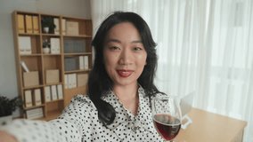 Happy young Asian woman with flute of red wine looking at camera in home environment while communicating in video chat, toasting and congratulating you on holiday