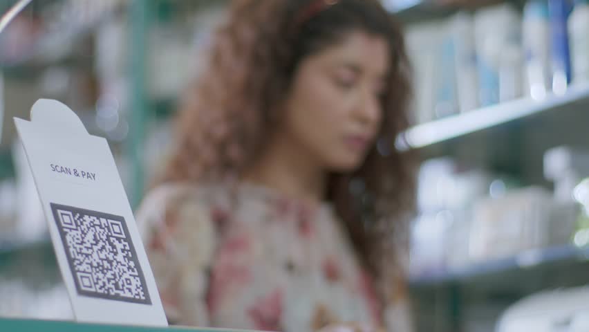 A close-up shot of a hand of a customer scanning a barcode to pay e-money to an electronic bank account or online UPI transfer in a general or grocery store with a smiling woman in the background Royalty-Free Stock Footage #1101265169