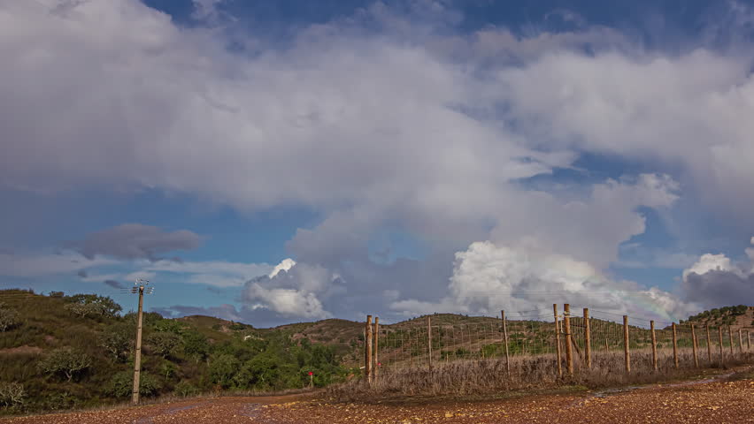 Blue sky with rainbows and white clouds moving over a dry field with fences. Take timelapse from below.