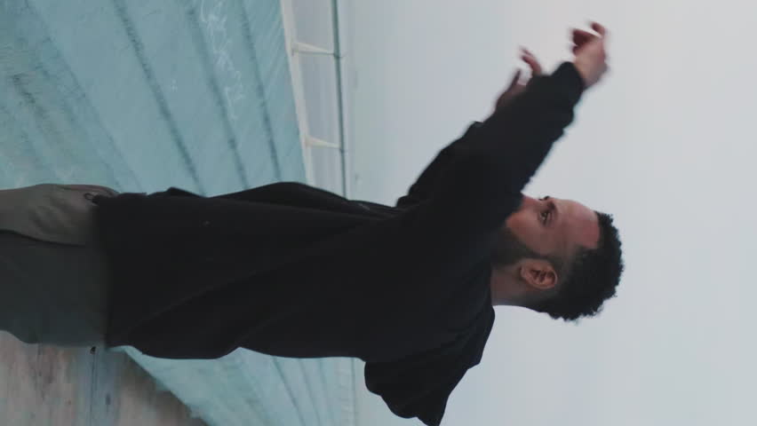 VERTICAL VIDEO: Young bearded male athlete doing warm-up, stretching his arms before training while standing on the embankment