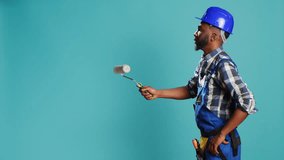 Industrial worker in uniform painting walls with roller, using renovation instrument over blue backdrop. Professional craftsman with hardhat working with rolling paint brush to change color. - Powered by Shutterstock - Get 15% off with code: PIKWIZARD15