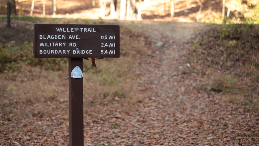 A signpost for the Valley Trail stands amidst the colorful foliage of Rock Creek Park in Washington, DC. Runners rush by in the foreground, capturing the essence of autumn.