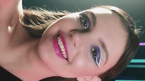 Vertical video. 90s makeup. Retro fashion. 2000s night look. Smiling confident teen girl face with blue color eyeshadow pink lips artistic visage posing in neon light on dark.