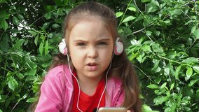 little girl with a phone and headphones outdoors in a garden or park. watching cartoons, playing games. modern kids and gadgets. Smiling girl listening music using smartphone wearing white headphones