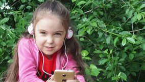 little girl with a phone and headphones outdoors in a garden or park. watching cartoons, playing games. modern kids and gadgets. Smiling girl listening music using smartphone wearing white headphones