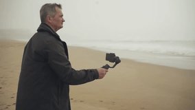 Senior caucasian man filming on the beach using a phone gimbal. Woman looking at the sea in the background