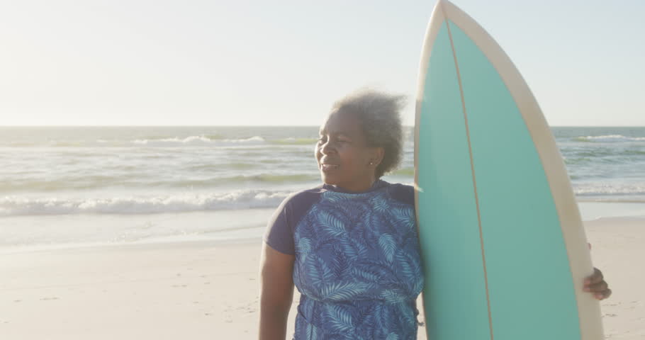 Happy senior african american woman holding surfboard at beach, in slow motion, with copy space. Spending quality time, lifestyle, retirement and vacation concept.