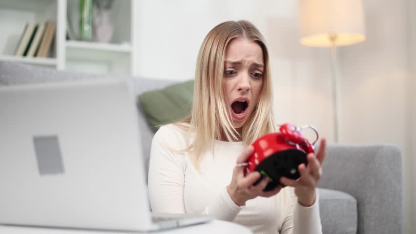 Nervous stressed young blond woman freelancer typing quickly on laptop computer and looking at clock in her hands asking to hurry up scream and warning about deadline at home