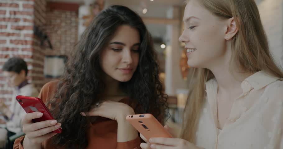 Portrait of young lady using smartphone showing screen to friend laughing having fun in cafe. Modern technology and social media concept.