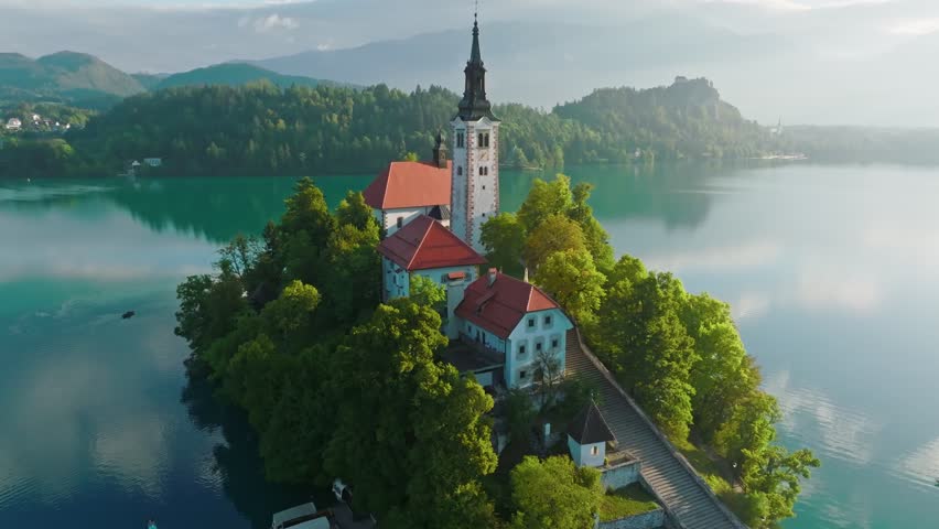 Flying around small island on Bled Lake in Slovenia. Aerial view of Church of the Assumption of Mary in the center of the lake Bled. Warm morning sun and light fog