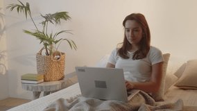 A female freelancer works remotely at a laptop while lying in bed. A young woman is lying in bed in the early morning typing something on a laptop.