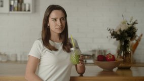 View of a cute smiling woman leaning on the table. Brunette woman smiling and looking at the camera holding cucumber juice in her hands.