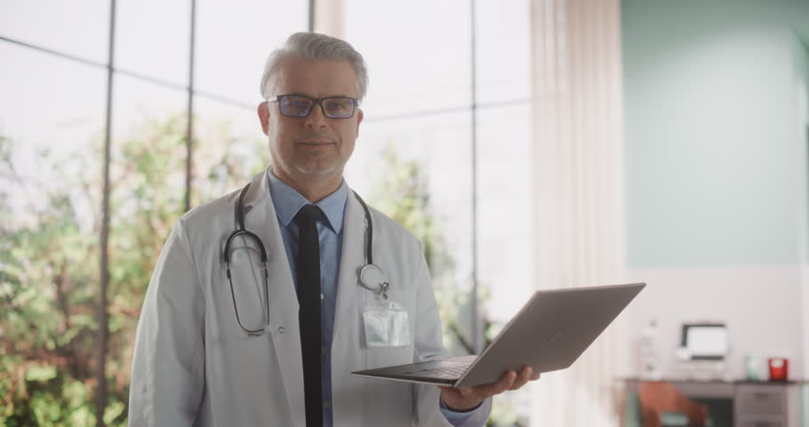 Portrait of an Experienced Middle Aged Handsome Doctor Wearing White Coat, Holding a Laptop Computer in the Clinic. Adult Medical Health Care Professional Looking at the Camera and Gently Smiling | Shutterstock HD Video #1101315663