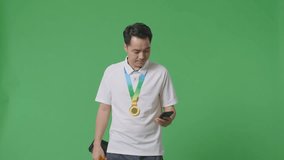 Asian Man With A Gold Medal Showing A Gold Trophy To The Video Call On Smartphone On Green Screen Background In The Studio
