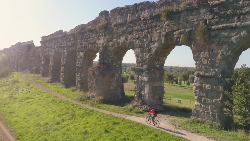Young man cyclist riding bike on dirt road along ancient roman aqueduct in orange sportswear and backpack aerial view drone dolly Royalty-Free Stock Footage #1101319135