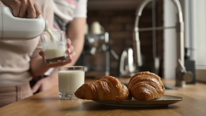 Girl pouring milk to glass and give to man for breakfast with croissants at kitchen. Woman and guy couple with white organic latte and french pastry Royalty-Free Stock Footage #1101319683