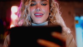 Female with natural make up and white teeth smile stands on night city street neons with mobile phone lighting her face. Millennial young woman using smartphone app, watching content in social media