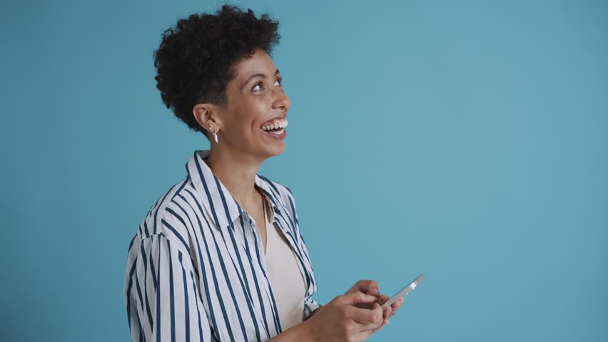 Laughing hispanic curly haired woman texting on phone while standing in the blue studio | Shutterstock HD Video #1101320843