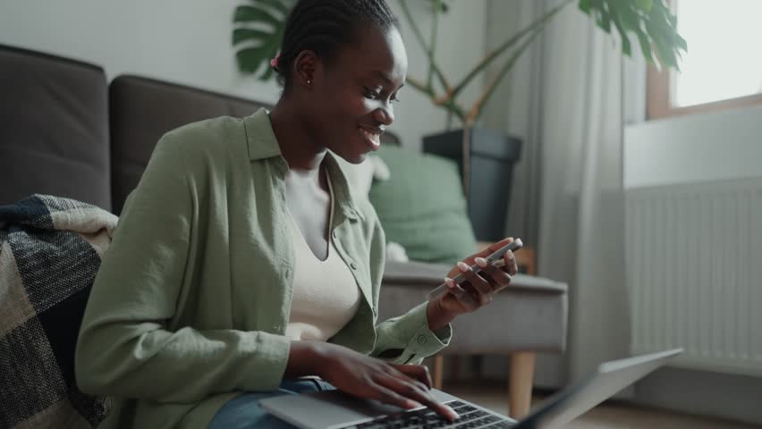 Positive African woman with pigtails typing something from a phone to a laptop while sitting on the floor at home