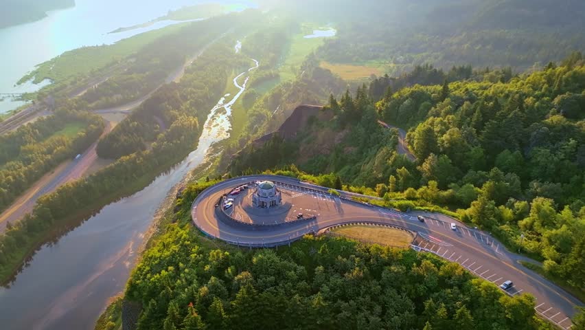 Aerial at golden hour during sunrise-a slow flying forward over and past Vista House showing Mirror Lake, the Columbia River and a train traveling East in the Columbia River Gorge-greens-blues-yellows