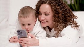 Mom and little 2-year-old son are lying on the bed, looking at a mobile phone, laughing, watching cartoons, making video calls, having fun on the Internet. Two generation family use new app