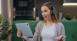 Pretty happy smiling caucasian young woman in headphones talking on video on tablet while sitting on sofa in room. Happy relaxed girl enjoys a conversation, gossip or chat with friend.