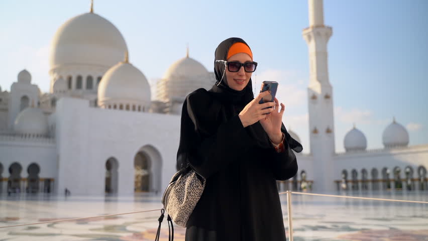 Woman in hijab has online call on background of mosque in UAE spbas. Young female holding smartphone in hand and looking at screen, talking with happy smile and standing near white Arabic architecture