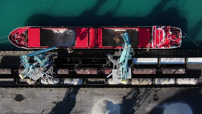 Port cranes unload a ship with mineral fertilizers into railway cars, aerial view. Transportation of goods by sea and rail.