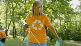 Caucasian Senior Female Eco Volunteer Using Trash Picker While Collecting Plastic in Bin Bag. Group of Eco Activists in T-Shirts with Recycling Symbol Cleaning Park. Safe Ecology Concept - Powered by Shutterstock - Get 15% off with code: PIKWIZARD15