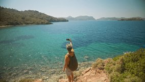 happy free man traveler with backpack makes selfie or video call standing on cliff on shore of beautiful sea bay with clear turquoise water and paradise empty sea beach. slow motion, travel in Turkey