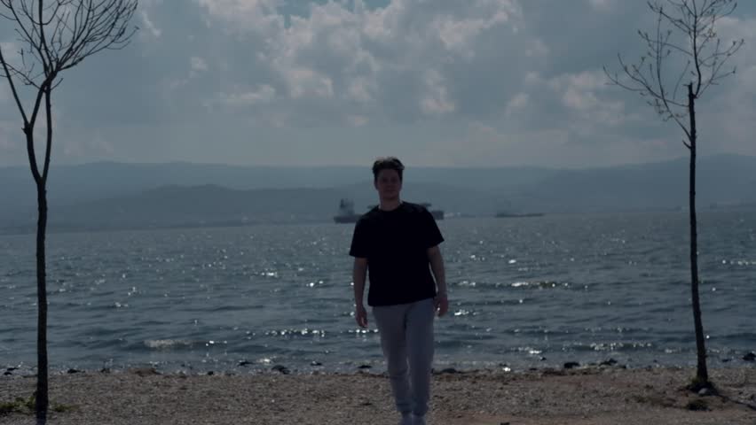 Teenager standing alone on the beach going, distant view of the ship standing in the middle of the sea, man coming towards the camera