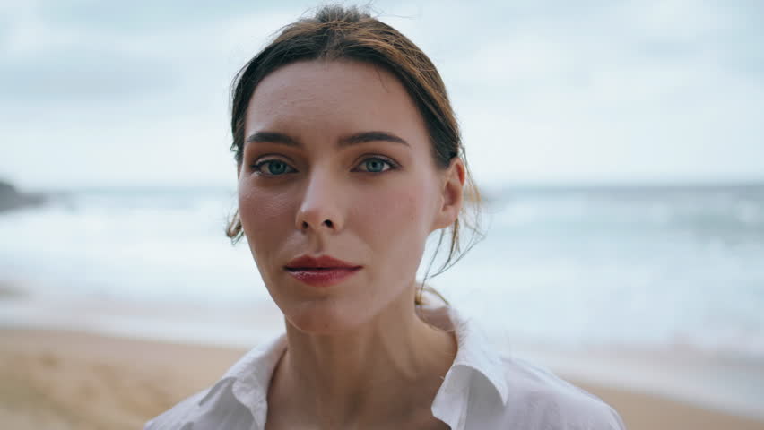 Beautiful gentle woman face looking camera intently in front sea waves closeup. Portrait of attractive pensive girl standing alone at cloudy beach wearing white shirt. Tranquil lady posing on seashore