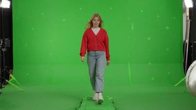 Attractive blond red-haired girl walks towards the camera in front of a green screen. Happy young woman in jeans and knitted jumper pointing with hands at advertising space. Chroma key. 4k UHD video