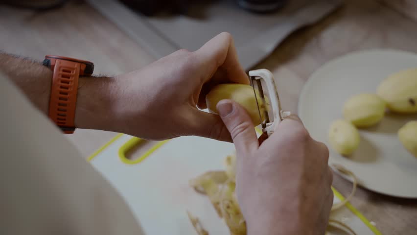 Caucasian male peeling potatoes over table in domestic kitchen. Cleaning potato. Cooking boiled pratie. Food preparation concept. Unrecognizable person holding knife and peel spud.